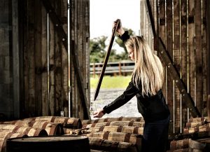 Woman in open barn uses a valinch (long, tapered copper tube) to extract whisky from a cask.